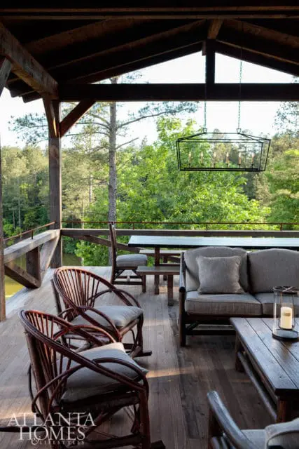 Covered porch with rustic wooden furniture, including rocking chairs, a sofa, and coffee table, overlooking a wooded landscape and water.