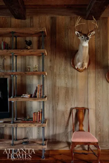 Rustic room with wood-paneled walls, a mounted deer head, a wooden chair with a pink upholstered seat, and a metal and wood bookshelf filled with books.