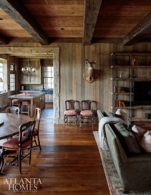 Rustic living room with exposed wooden beams, wood-paneled walls, a mounted deer head, and a large sectional sofa. An open doorway reveals a kitchen with a central island and pendant lighting.