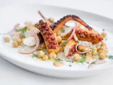 Grilled octopus tentacles served with chickpeas, thinly sliced red onion, and fresh herbs on a white oval plate.