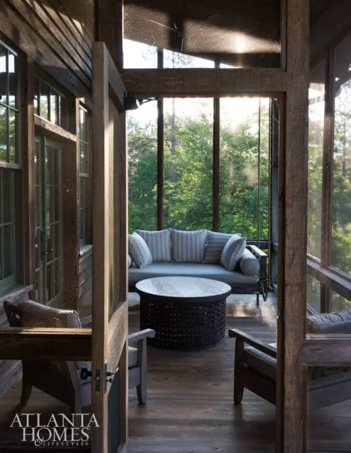 Screened porch with rustic wood beams, a gray sofa with striped pillows, a dark metal coffee table, and two wooden chairs. Sunlight streams through the windows, illuminating trees outside.