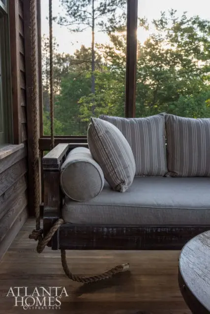 Screened porch with a wooden swing couch featuring grey cushions and striped pillows, overlooking trees at sunset. Atlanta Homes & Lifestyles logo in the corner.