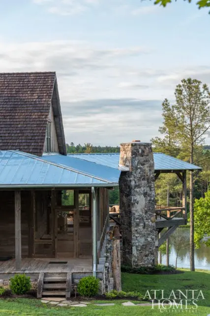 Rustic wooden house with a stone chimney, metal roof, and screened-in porch overlooking a lake and wooded area.