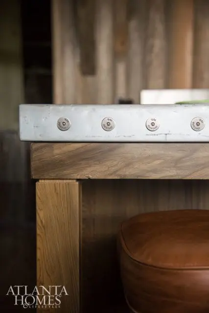 Close-up of a light gray countertop with four decorative metal discs, each labeled '12 GA BUCKSHOT', set into the surface above a wooden base and a brown leather stool.