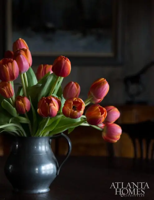 Close-up of orange and pink tulips in a pewter pitcher on a dark wood table, with the Atlanta Homes & Lifestyles logo in the lower right corner.