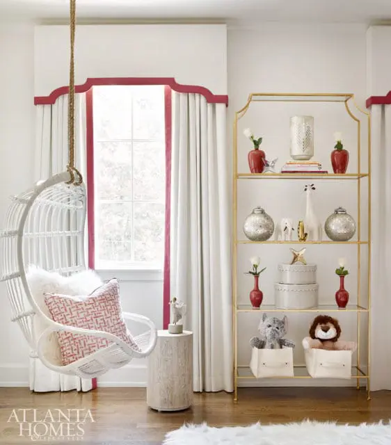 White hanging chair with pink patterned pillow next to a wood side table and a gold shelf displaying decorative items and stuffed animals.
