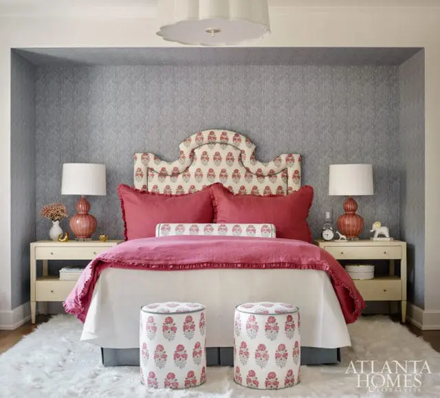 Bedroom with a gray patterned wallpapered alcove, a white bed with pink and red floral headboard, red pillows, and two white nightstands with coral lamps.
