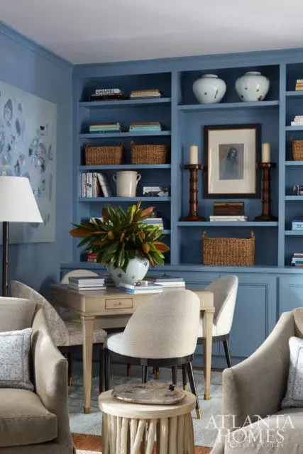Blue built-in bookshelves filled with books, baskets, and decorative vases. A light wood desk with chairs sits in front, adorned with a large floral arrangement.