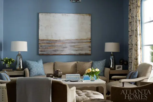 Living room with blue walls, a large abstract painting, beige sofa with blue patterned pillows, and a white coffee table with floral arrangement.