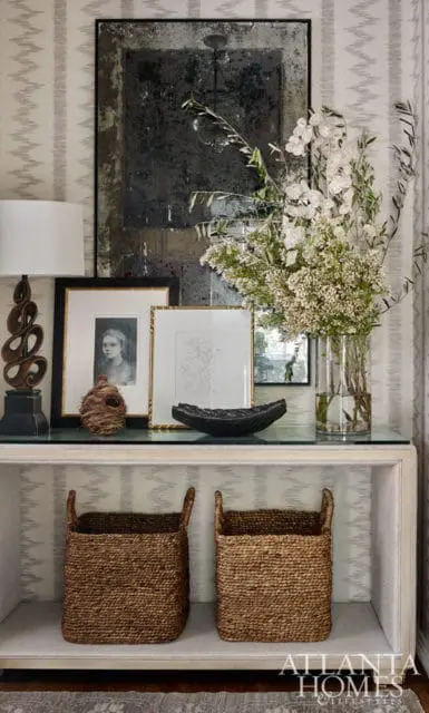 Console table with a glass top displaying framed artwork, a vase of white flowers, and decorative objects. Two woven baskets sit on the shelf below.