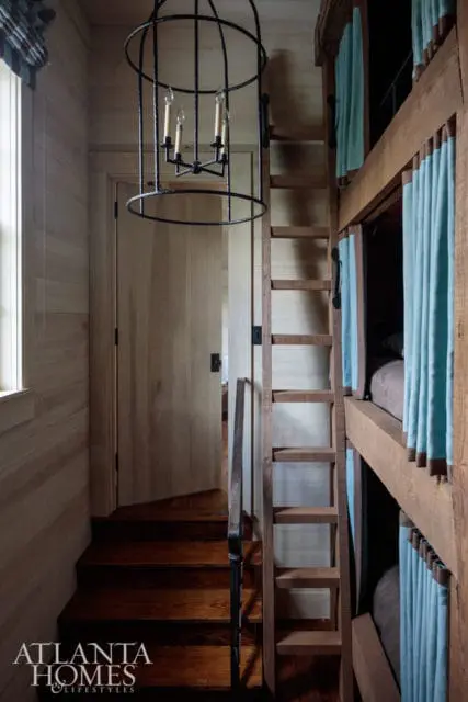 Hallway with wood-paneled walls, a wooden door, a dark metal chandelier, and built-in bunk beds with light blue curtains. A wooden ladder leads to the upper bunks.