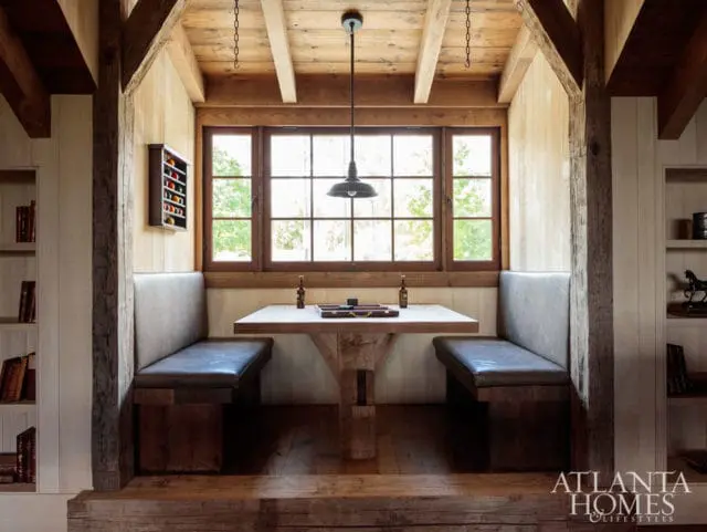 Rustic dining nook with built-in upholstered benches, a rough-hewn wood table, and a large window overlooking trees. Exposed wood beams and paneled walls create a cozy atmosphere.