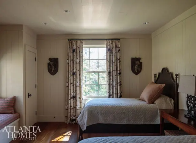 Bedroom with two twin beds, paneled walls, wood floors, and a window with patterned curtains showing trees outside.