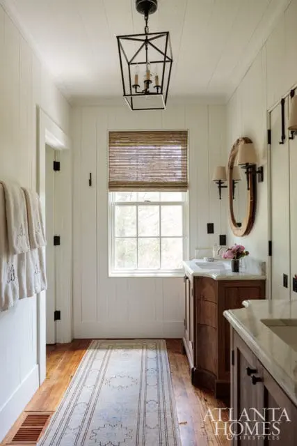 Narrow bathroom with white shiplap walls, a wooden vanity with a marble countertop, a round mirror, and a geometric lantern pendant light.