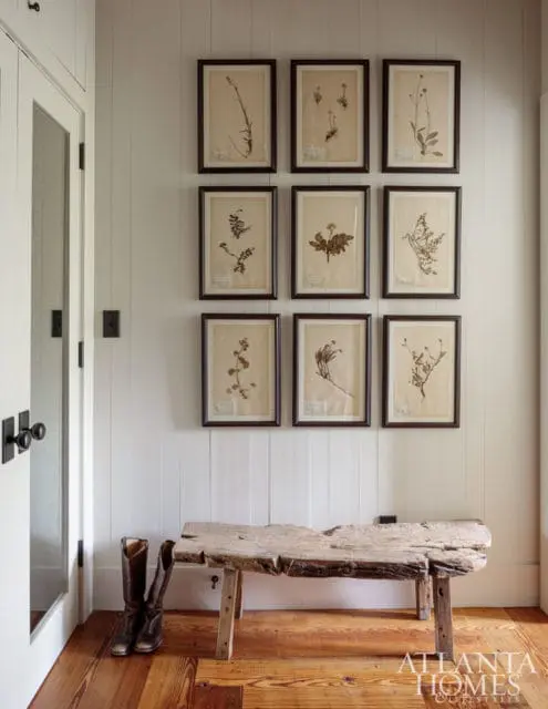 Hallway with white paneled walls displaying nine framed botanical prints, a rustic wooden bench, and a pair of cowboy boots on the wooden floor.