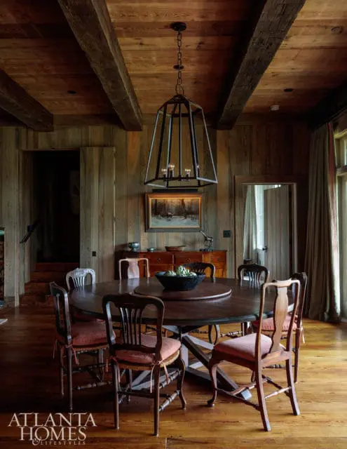 Rustic dining room with dark wood paneled walls, beamed ceiling, round wooden table with upholstered chairs, and a large geometric chandelier.