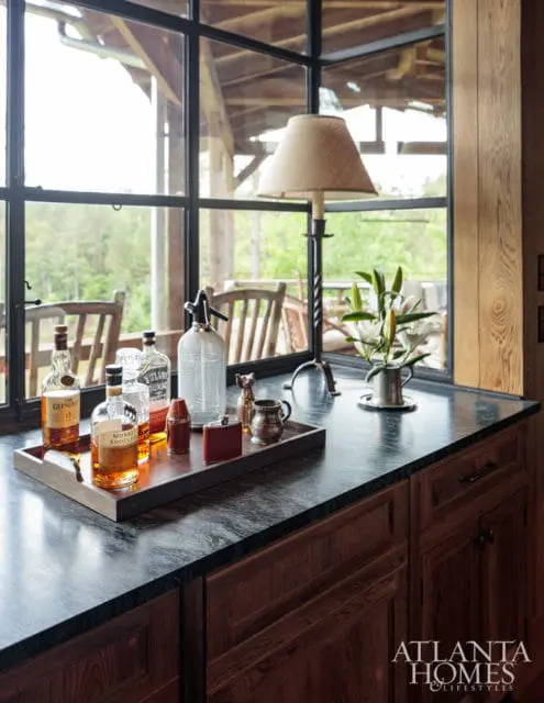 Dark granite countertop with a wooden tray holding bottles of whiskey, a soda siphon, and bar accessories. A lamp and vase of lilies sit behind the tray.