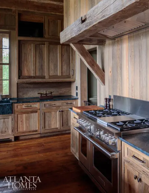 Rustic kitchen with natural wood cabinetry, dark stone countertops, stainless steel range, and exposed wooden beam above the range hood.