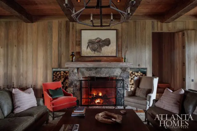 Rustic living room with wood-paneled walls, exposed beams, and a stone fireplace with a painting of a turkey above. Features include a red armchair, leather sofas, and a dark wood coffee table.