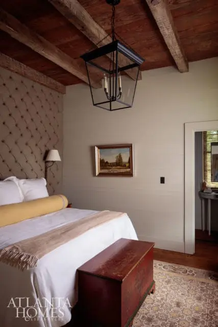 Bedroom with tufted headboard, white bedding, yellow bolster pillow, and a dark wood ceiling with exposed beams. A black lantern hangs overhead.