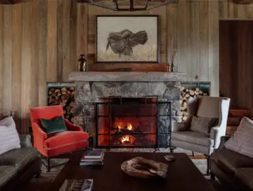 Living room with wood-paneled walls, stone fireplace with a painting of a turkey above, and comfortable seating including a red armchair and leather sofas.