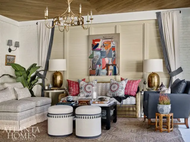 Living room with a sectional sofa, patterned pillows, two ottomans, a coffee table, and a gold chandelier. The walls are paneled and brick, with a slatted blind behind the sofa.