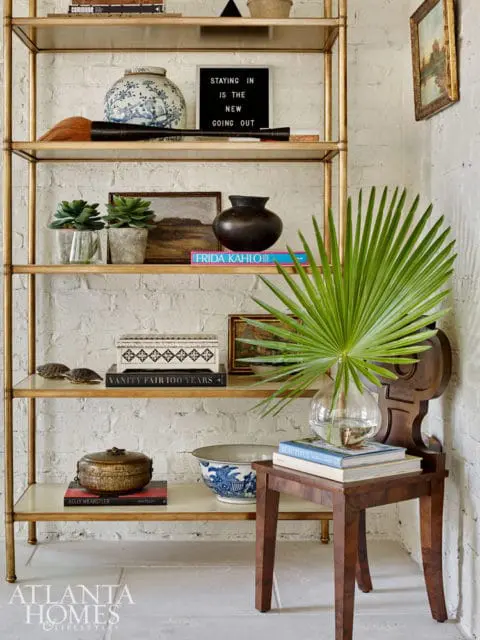 Gold etagere filled with decorative objects including vases, books, plants, and a framed painting against a white brick wall.