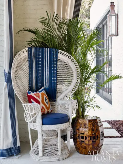 White wicker peacock chair with blue cushion and patterned pillows, next to a large potted palm and a decorative metal garden stool. Blue and white striped curtains hang to the left.