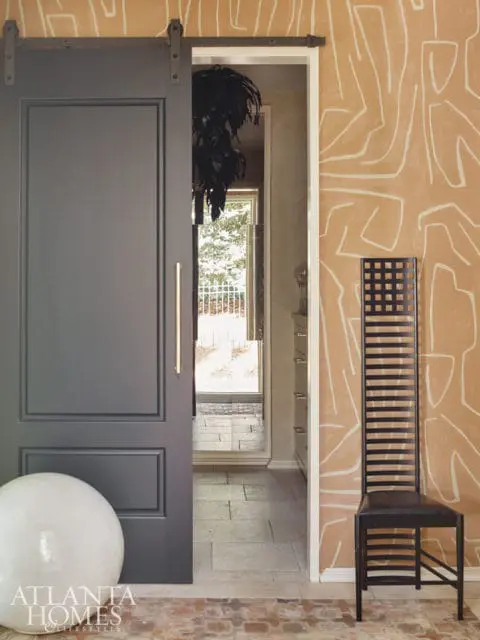 Dark gray sliding barn door with brass handle next to tan wallpaper with white abstract lines. A black metal chair with a tall slatted back sits in the corner.