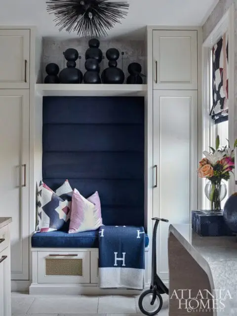 Built-in banquette with navy blue tufted upholstery, geometric and watercolor pillows, and a navy blanket with white 'H' logos. Above, a shelf displays black abstract sculptures and a spiky light fixture.