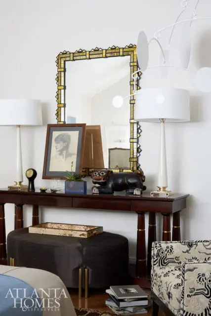 Dark wood console table topped with a framed portrait, a black carved animal sculpture, and two white lamps. A dark upholstered ottoman sits below the table.