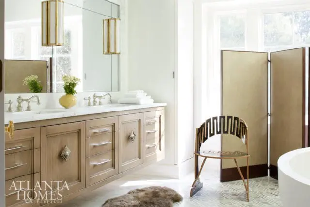 Modern bathroom with a light wood double vanity, marble countertop, silver faucets, and two cylindrical brass and glass pendant lights. A decorative chair and a folding screen are visible near a bathtub.