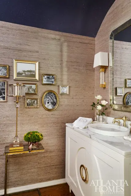 Powder room with grasscloth wallpaper, a navy speckled ceiling, a white vanity with marble countertop, and a collection of framed black and white photographs on the wall.