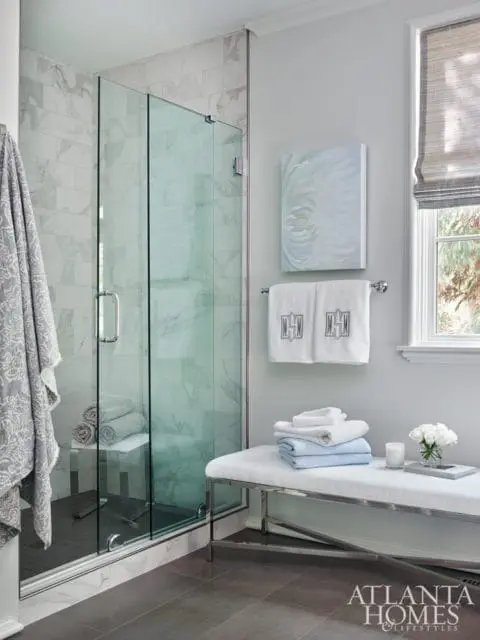 Modern bathroom with glass-enclosed shower featuring marble tile, a white upholstered bench with stacked towels, and a window with a roman shade.