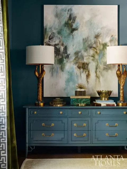 Teal dresser with gold hardware topped with books, a decorative bowl, and two brass lamps with bird bases, beneath a large abstract painting.