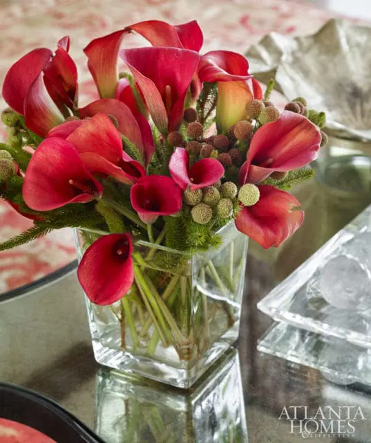 Close-up of a bouquet of red calla lilies and green foliage in a square glass vase on a reflective surface.