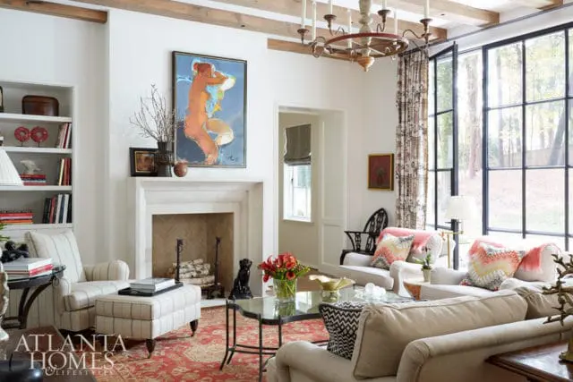 Living room with white walls, exposed wooden beams, a stone fireplace, and a large abstract painting above the mantle. Furniture includes striped armchairs, sofas with patterned pillows, and a glass coffee table.