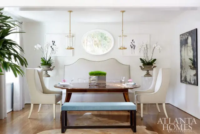 Dining room with oval wood table, upholstered chairs, and a light blue bench. A large upholstered headboard is behind the table, with two gold pendant lights overhead.