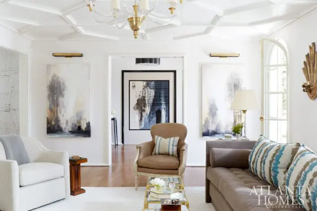 Living room with white walls, coffered ceiling, abstract art, tufted sofa, leather armchair, and glass coffee table.