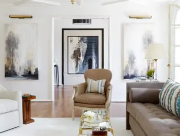 Living room with tufted taupe sofa, leather armchair, and white sofa, featuring abstract art and a mirrored coffee table.