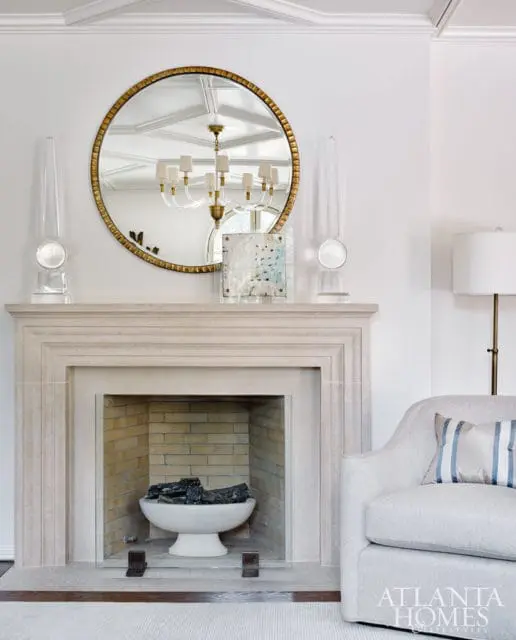 Limestone fireplace with a gold-framed round mirror above, flanked by clear obelisk sculptures. A white armchair with a striped pillow sits to the right.