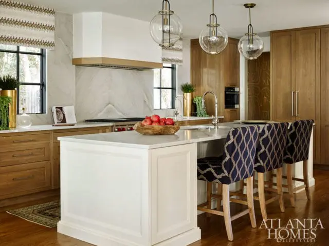 Kitchen island with white cabinetry and marble countertop, featuring upholstered bar stools with a navy and gold geometric pattern. Natural wood cabinets and marble backsplash are visible.
