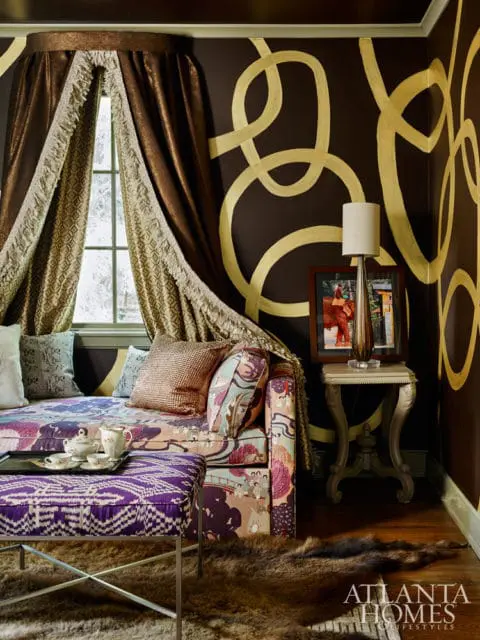 Dark brown walls with gold abstract swirls, a patterned daybed with colorful pillows, a purple patterned ottoman, and metallic curtains framing a window.