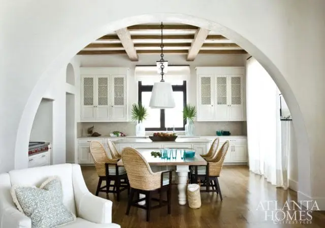 White kitchen with beamed ceiling, white cabinetry with lattice doors, and a dining table with woven chairs.