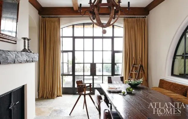 Dining room with dark wood table, stone floor, large arched window with metal panes, and gold drapes.