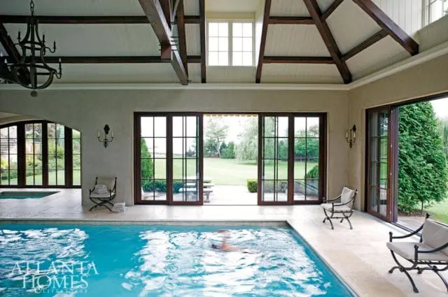 Indoor swimming pool with vaulted wood beam ceiling, large glass doors opening to a manicured lawn, and a person swimming.