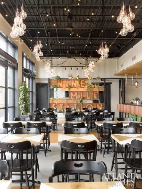 Restaurant dining room with black chairs and light wood tables, featuring a large wooden sign with 'NINE MILE STATION' and hanging Edison bulb light fixtures.