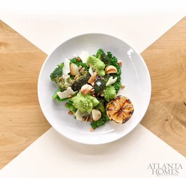 Broccoli and Romanesco salad with shaved parmesan, toasted nuts, garlic slices, and a grilled lemon wedge on a white plate.