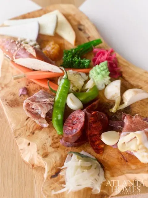 Charcuterie board with sliced meats, cheeses, pretzel, peppers, garlic, and pickled vegetables on a live-edge wooden board.