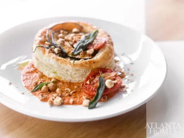Savory tart with flaky pastry, layered eggplant, tomatoes, and herbs, served with a red sauce and topped with chopped nuts on a white plate.